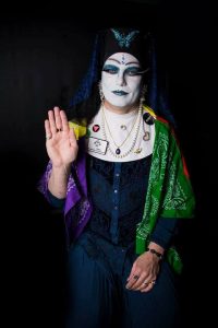 A person wearing white makeup and a nun's habit holds their hand up in a blessing gesture.