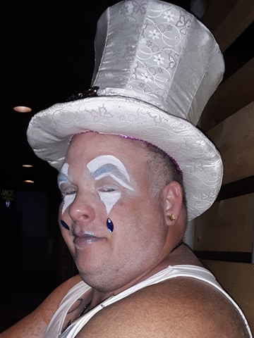 A close up of a person's head with white makeup and blue jewels attached to the face. They are wearing a white top hat