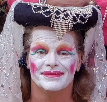 A person wearing white makeup with bright red lips, rouge, and eye makeup looks into the camera and smiles. They are wearing a black headress with a white veil on the back and a large rhinestone ornament in front